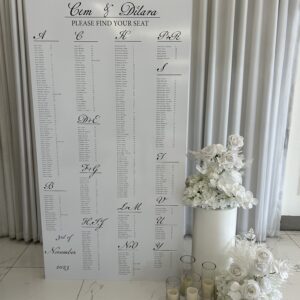 Modern white wedding seating chart board displayed with white plinths and glass candles.