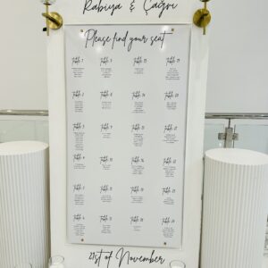 White framed seating chart display with gold and crystal accents, styled with white ripple plinths and candles for wedding décor.